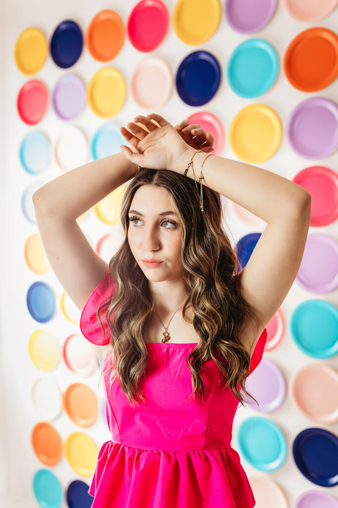 Teen girl in pink top posing with hands above head against colorful wall of plates.