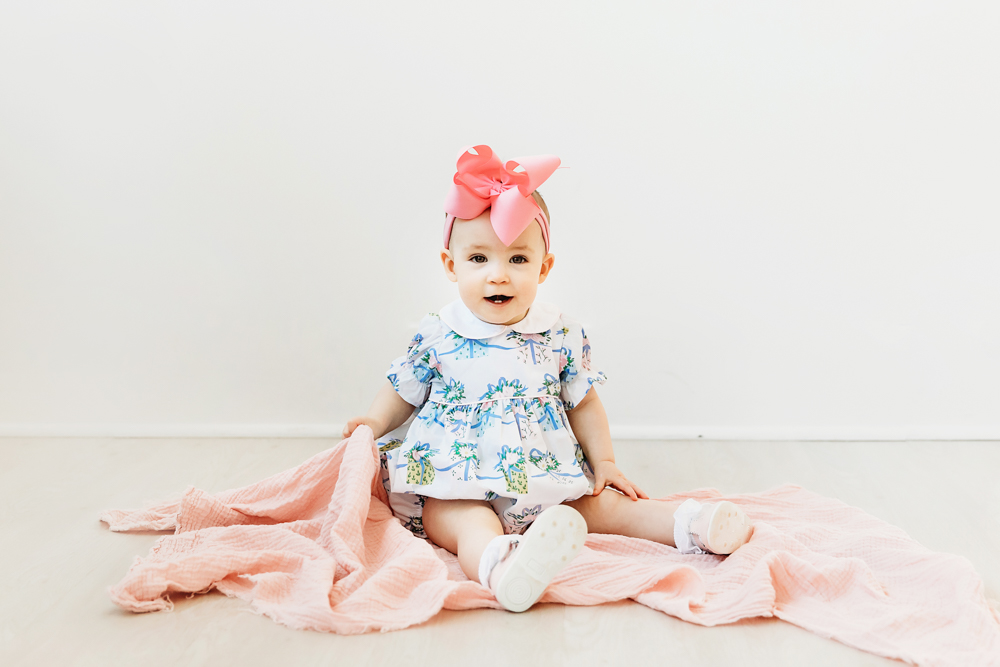 Elsie sitting on light floor with pink blanket, floral romper and oversized pink bow, milestone portrait at Reflection Images by Tracy Rowell in Florence SC