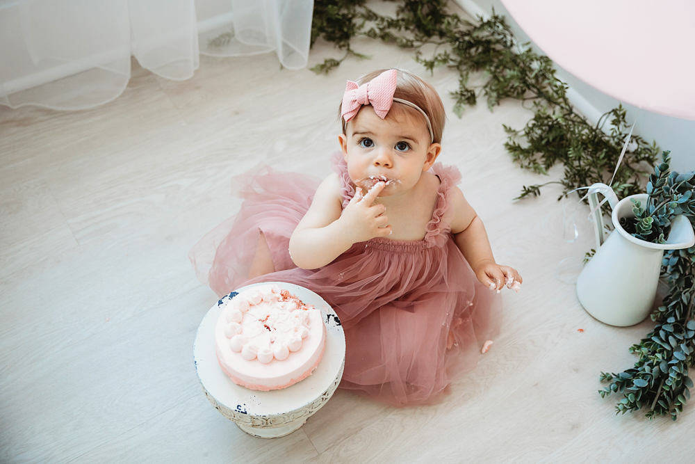 Grace enjoying a bite of pink smash cake in a ruffled mauve dress and bow, seated on light wood floor at Reflection Images by Tracy Rowell in Florence SC