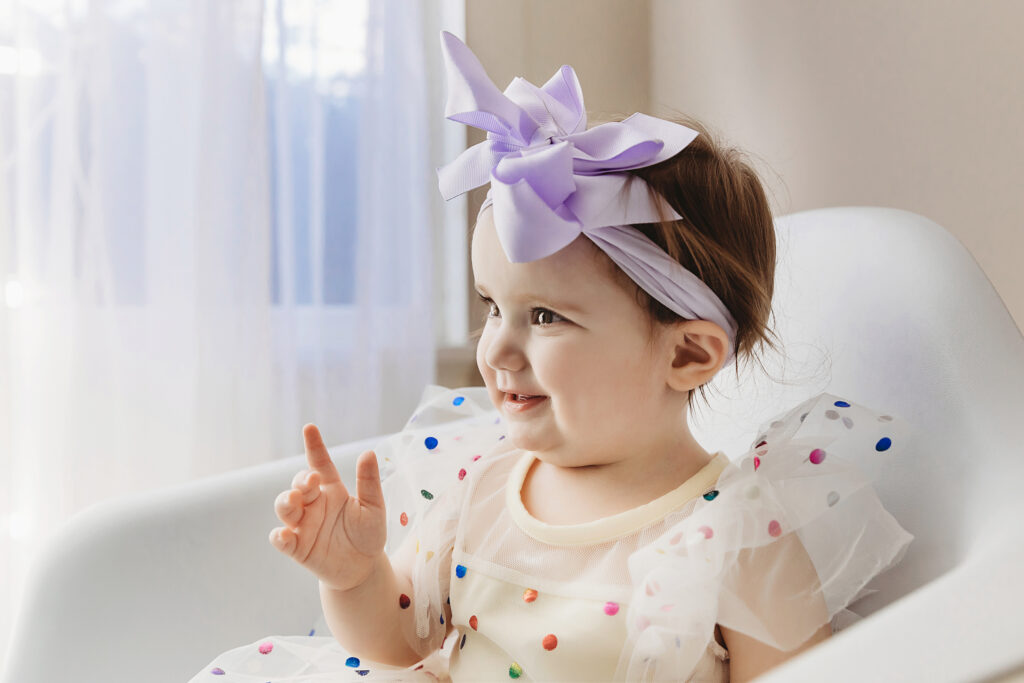 Side profile of Sullivan in a rainbow dress and lavender bow with soft window light coming through sheer curtains at Reflection Images by Tracy Rowell in Florence SC.