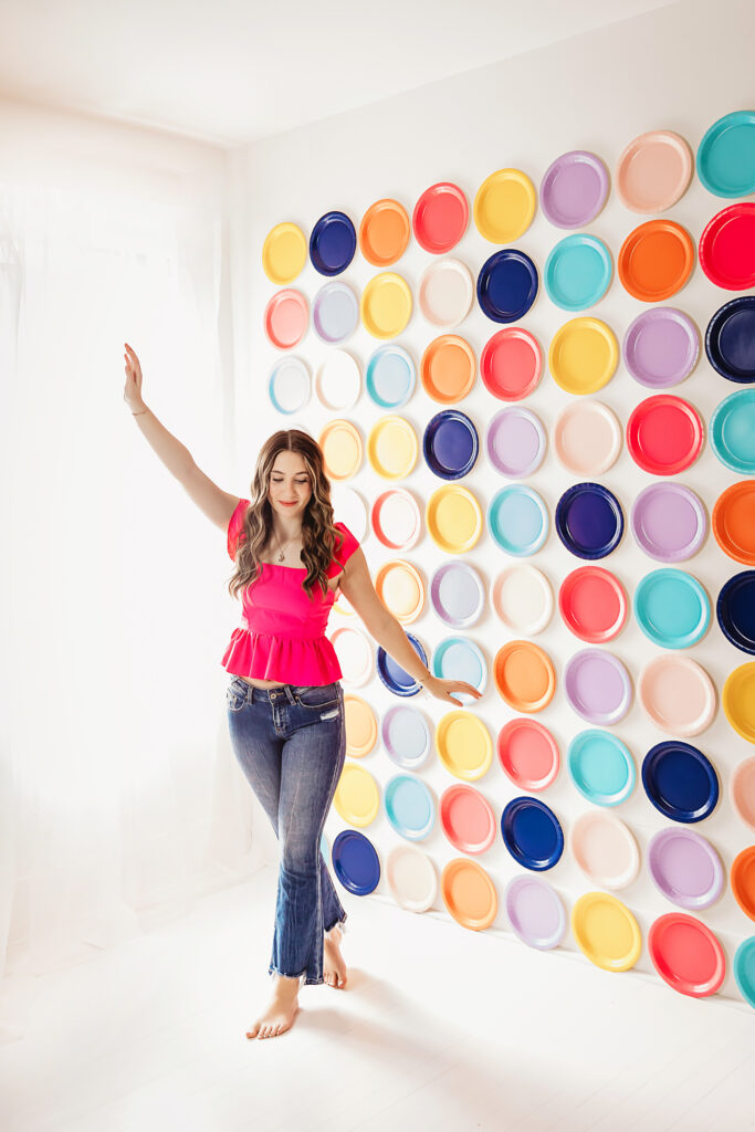 Teen girl in a bright pink top dancing barefoot in front of a rainbow wall of plates.