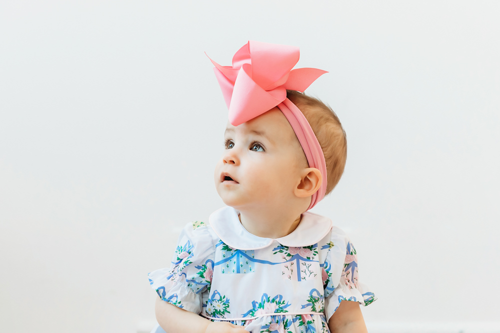 Elsie looking upward with curious gaze, floral romper, oversized pink bow, studio light backdrop at Reflection Images by Tracy Rowell in Florence SC