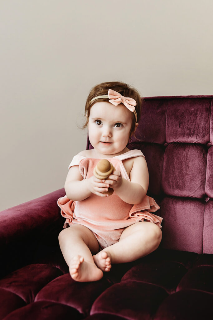 Sullivan in a blush velvet romper with a matching bow headband, seated on a plum velvet chair holding a wooden toy at Reflection Images by Tracy Rowell in Florence SC.
