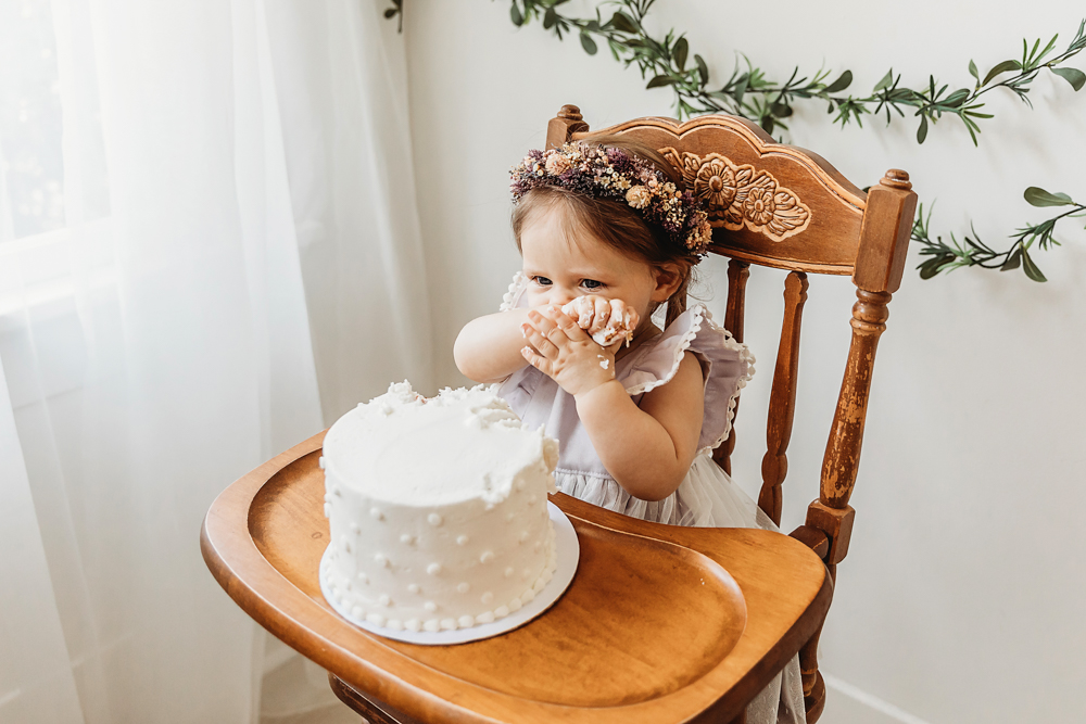 Baby girl covered in white cake icing while enjoying her first taste, sitting in a wooden high chair.