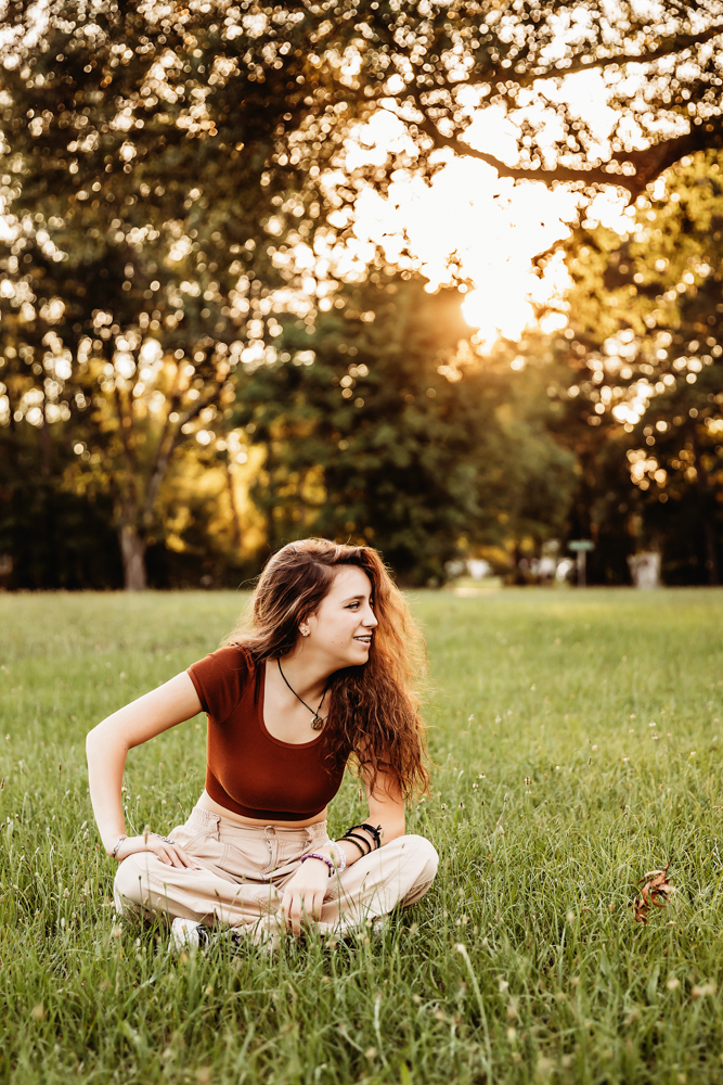 Eve sitting cross-legged in khaki pants and rust top, smiling with flowing hair in evening light at Reflection Images by Tracy Rowell in Florence SC