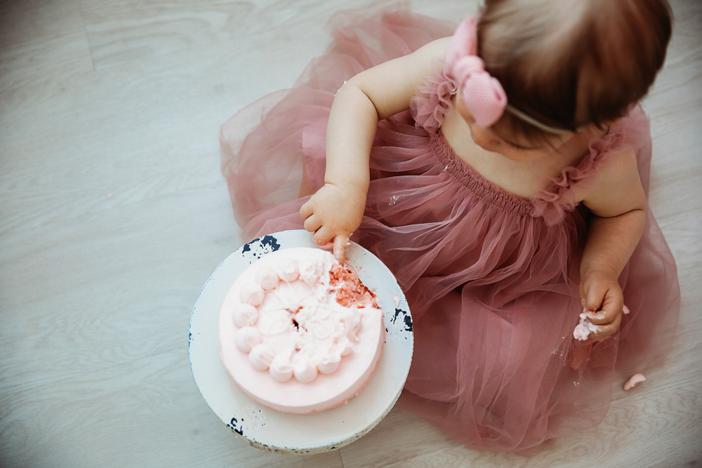 Top-down view of Grace digging into her pink cake while sitting on the floor in a tulle dress and headband at Reflection Images by Tracy Rowell in Florence SC