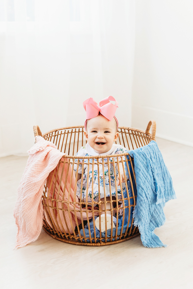 Elsie smiling in wicker basket, pink and blue textured blankets, floral romper with pink bow at Reflection Images by Tracy Rowell in Florence SC