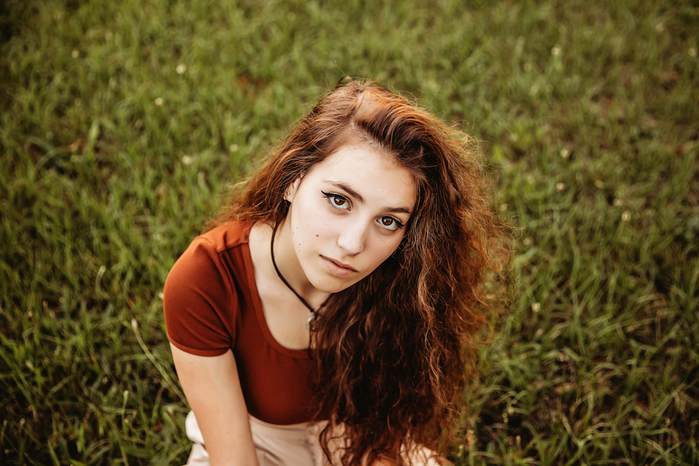 Close-up of Eve sitting in grass wearing rust top and khaki pants, direct gaze, soft natural light at Reflection Images by Tracy Rowell in Florence SC