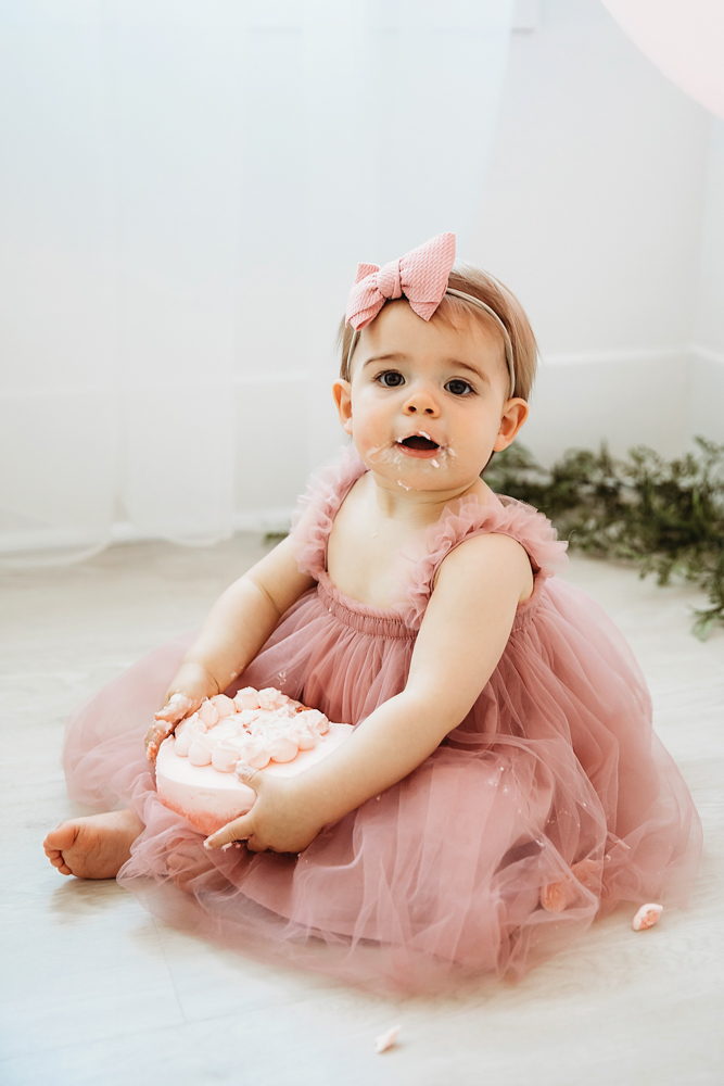 Grace holding the entire pink smash cake in her lap while seated near sheer white curtains and greenery at Reflection Images by Tracy Rowell in Florence SC