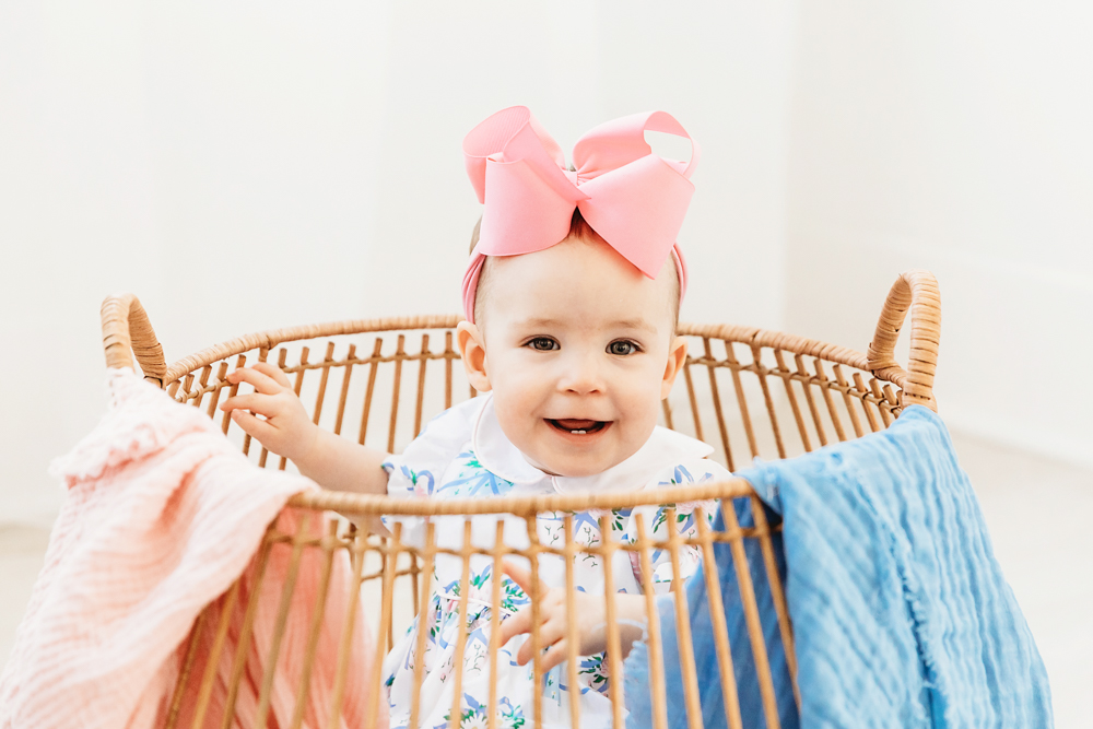 Close-up of Elsie inside wicker basket, floral romper with pink bow, smiling against soft window light at Reflection Images by Tracy Rowell in Florence SC