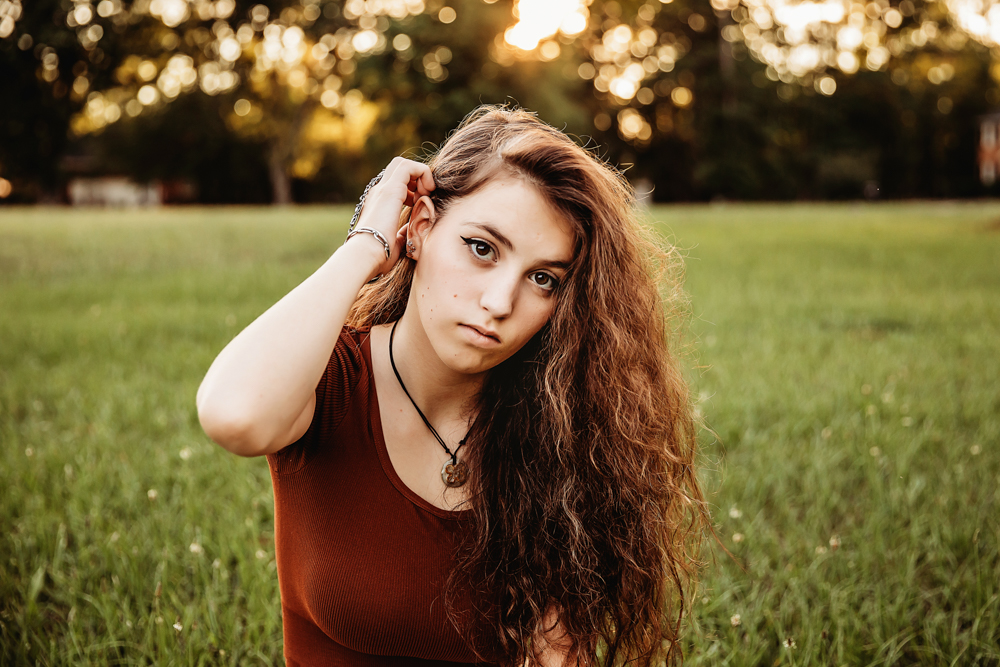 Eve sitting in grass with rust crop top, one hand touching hair, neutral expression, golden bokeh at Reflection Images by Tracy Rowell in Florence SC