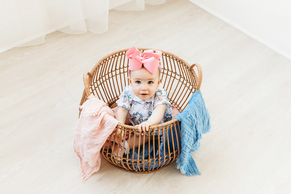 Elsie sitting in wicker basket with pink and blue blankets, floral romper, big pink bow at Reflection Images by Tracy Rowell in Florence SC