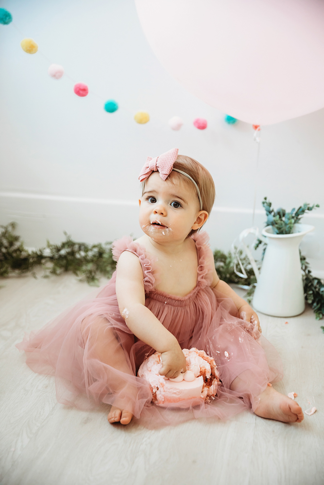 Grace surrounded by pink balloon and colorful felt garland, sitting with her cake in a mauve birthday dress at Reflection Images by Tracy Rowell in Florence SC