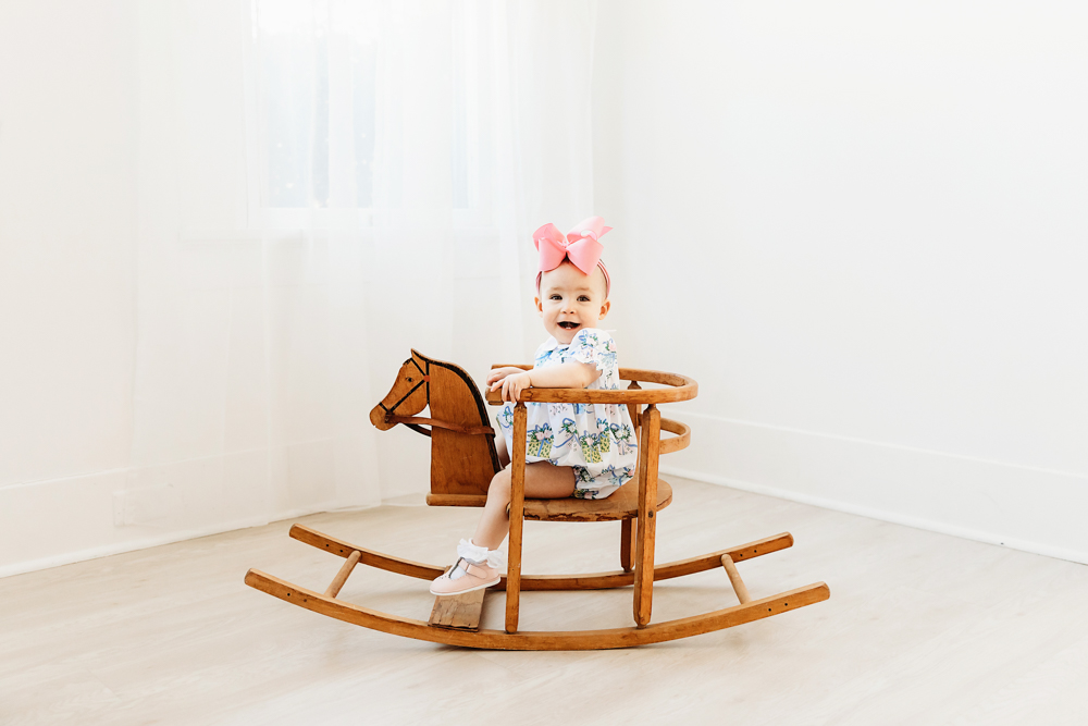 Elsie riding vintage wooden rocking horse, floral romper and oversized pink bow, natural light studio at Reflection Images by Tracy Rowell in Florence SC