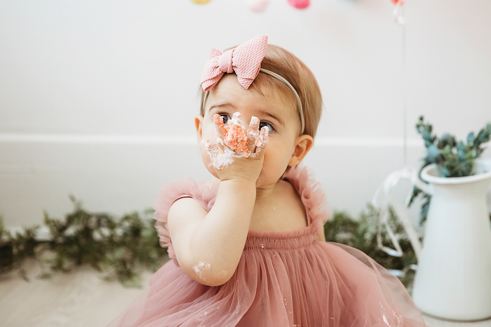 Close-up of Grace with a hand full of frosting held up to her mouth, colorful garland in background at Reflection Images by Tracy Rowell in Florence SC