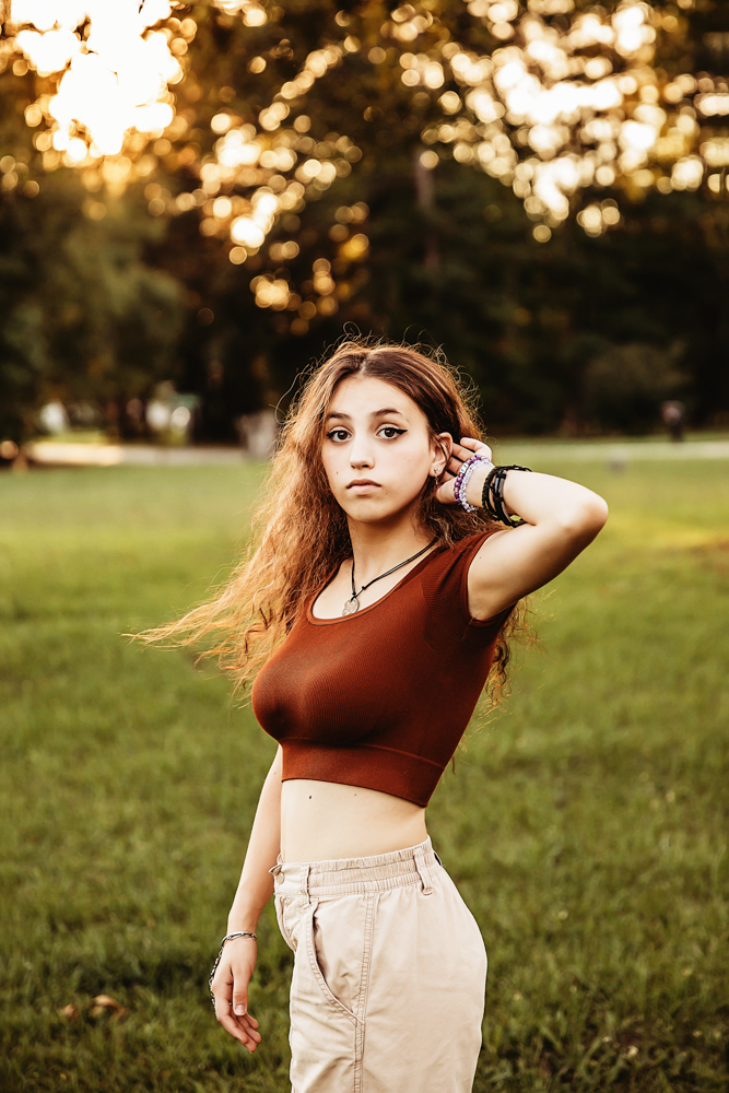 Eve standing in khaki pants and rust top, one hand brushing hair back, warm evening glow in open grassy field at Reflection Images by Tracy Rowell in Florence SC