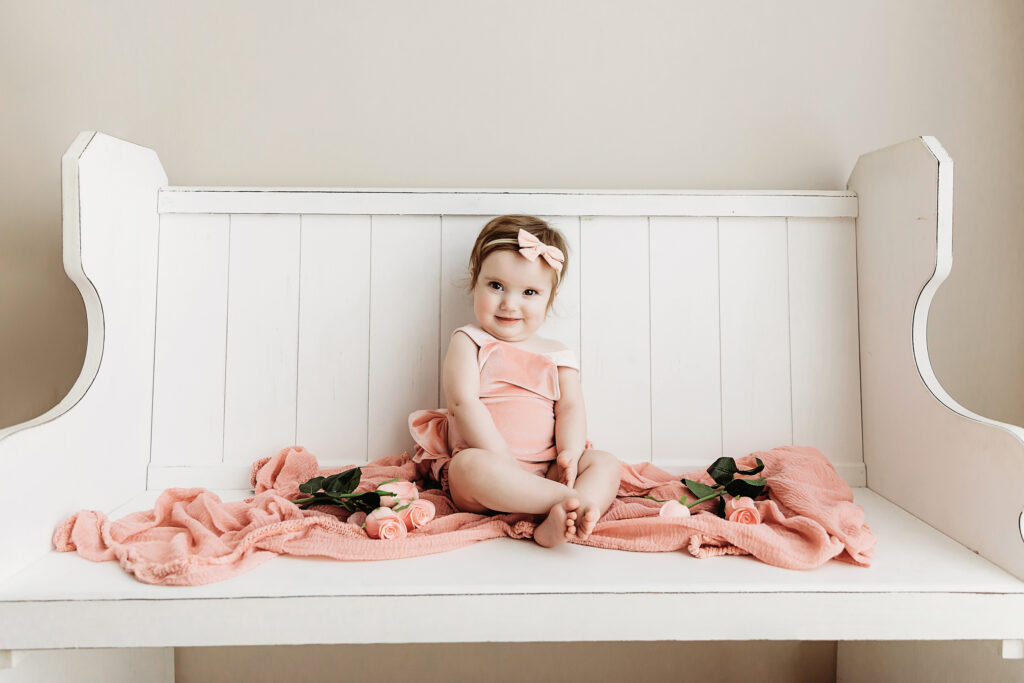 Sullivan in a blush romper on a white bench surrounded by blush roses and soft pink fabric at Reflection Images by Tracy Rowell in Florence SC.