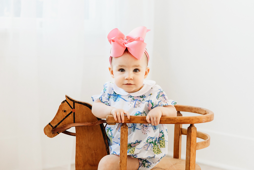 Close-up of Elsie on wooden rocking horse, floral romper with pink bow, soft window light at Reflection Images by Tracy Rowell in Florence SC
