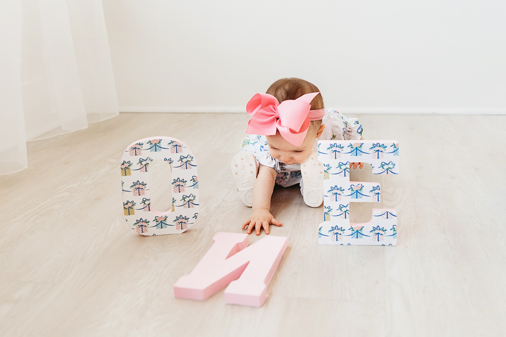 Elsie crawling between decorated wooden ONE letters, floral romper and pink bow, milestone setup at Reflection Images by Tracy Rowell in Florence SC