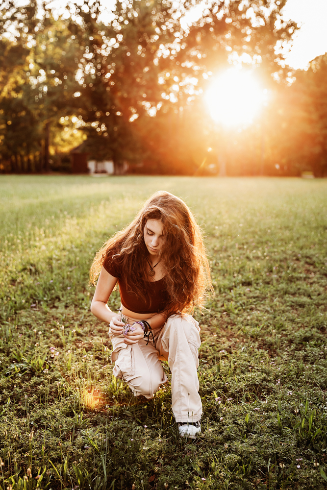 Eve kneeling in open grassy field, picking small flowers, rust crop top, khaki pants, dramatic sunset glow at Reflection Images by Tracy Rowell in Florence SC