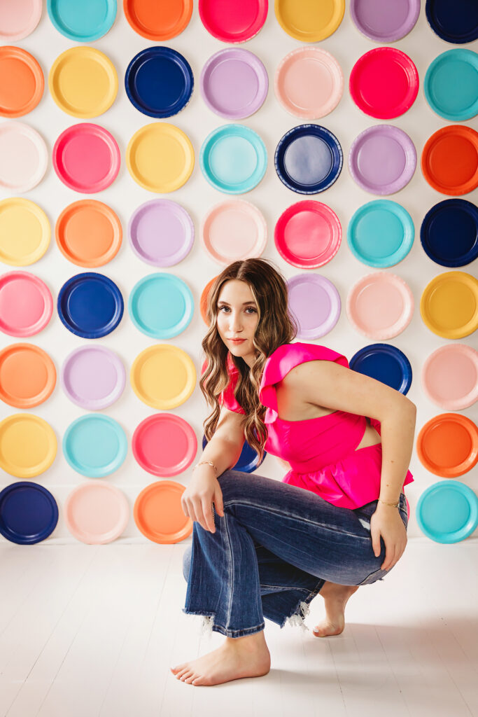 Teen girl squatting barefoot in front of a rainbow wall with a confident gaze.