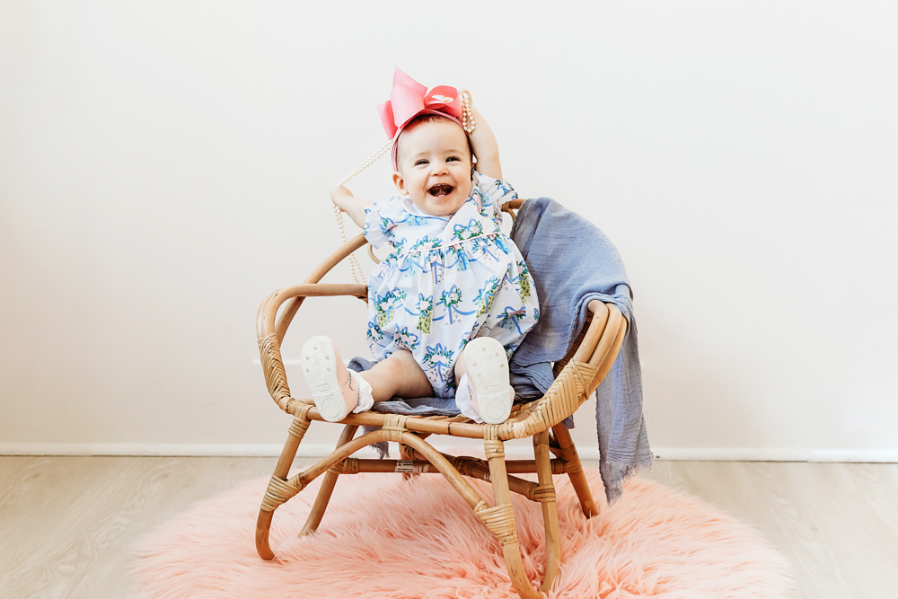 Elsie laughing while holding pearl necklace, floral romper with pink bow, sitting in wicker chair on pink rug at Reflection Images by Tracy Rowell in Florence SC