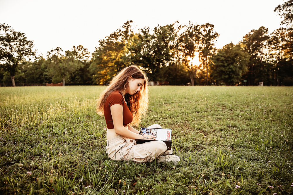 Eve seated in open grassy field with rust crop top, khaki pants, holding a magazine, soft golden sunset in background at Reflection Images by Tracy Rowell in Florence SC