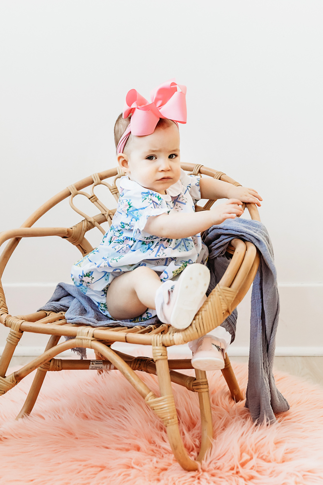 Elsie sitting in wicker chair on pink rug, floral romper with oversized pink bow, candid milestone at Reflection Images by Tracy Rowell in Florence SC