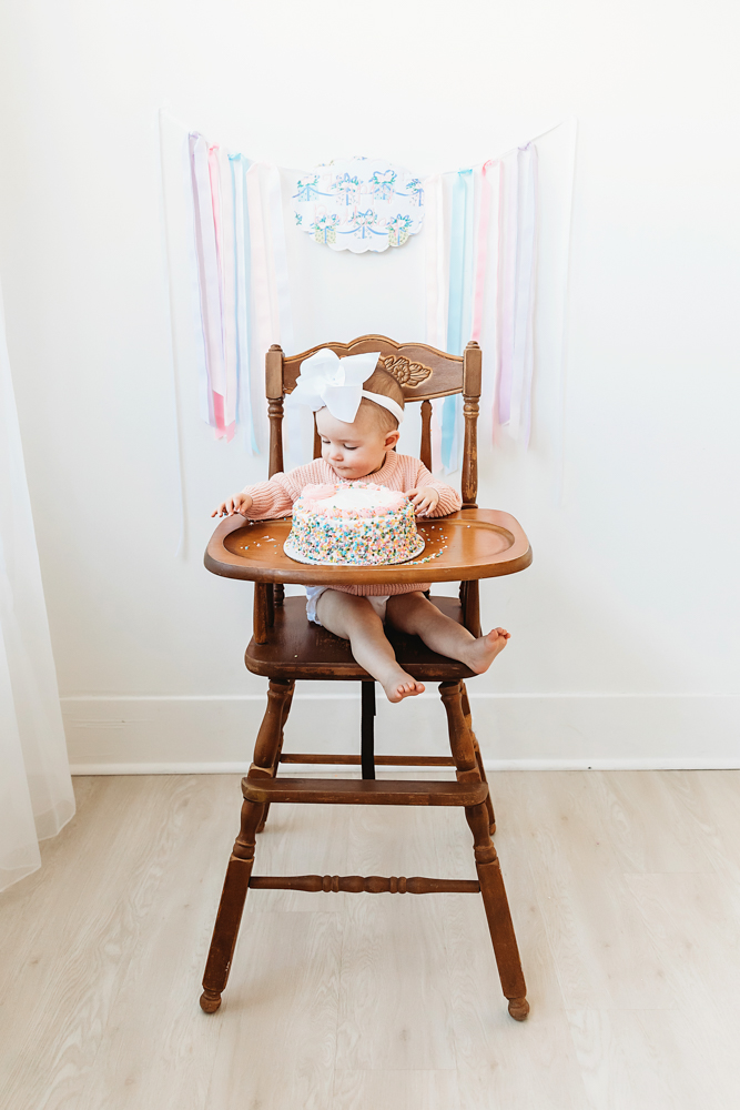 Elsie in wooden highchair with rainbow sprinkle cake, white bow and pink sweater, birthday banner backdrop at Reflection Images by Tracy Rowell in Florence SC