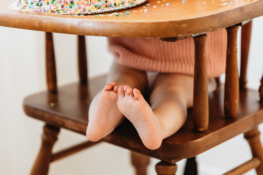 Close-up of Elsie’s tiny feet dangling under wooden highchair, birthday cake above, milestone detail at Reflection Images by Tracy Rowell in Florence SC
