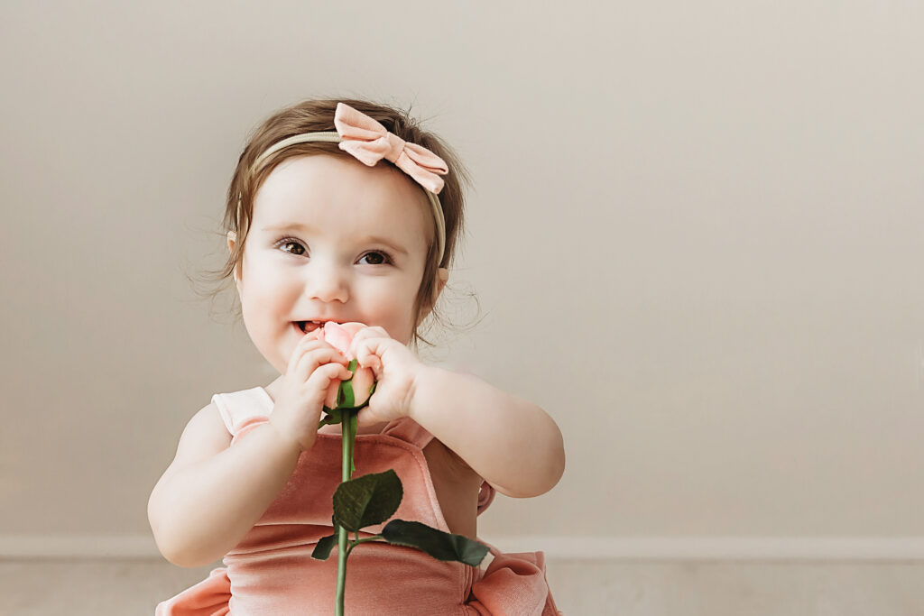 Sullivan smiling and holding a rose, wearing a blush bow and romper in front of a cream wall at Reflection Images by Tracy Rowell in Florence SC.