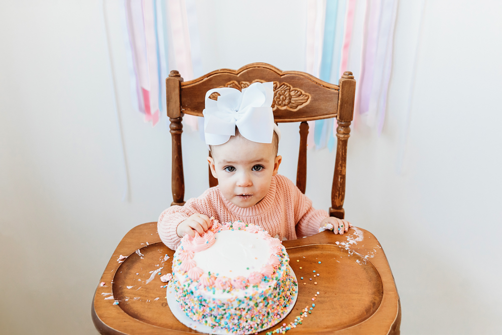 Elsie in pink sweater with white bow, tasting sprinkle birthday cake in wooden highchair, playful smash moment at Reflection Images by Tracy Rowell in Florence SC