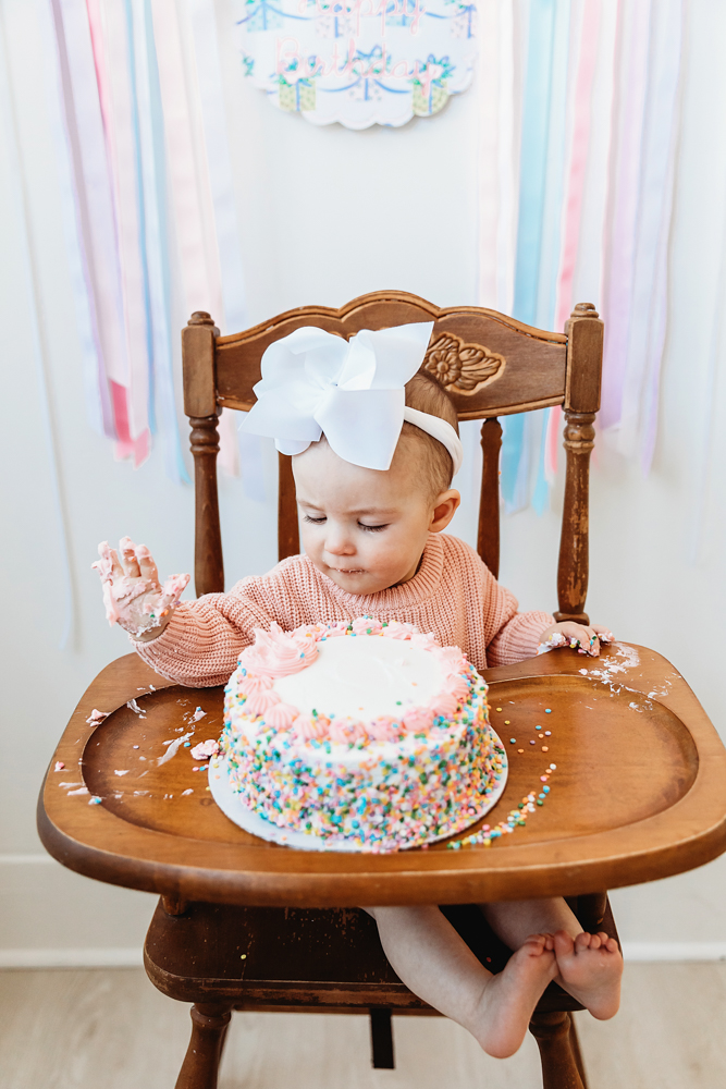 Elsie reaching into sprinkle-covered cake, pink sweater and white bow, cake smash milestone at Reflection Images by Tracy Rowell in Florence SC