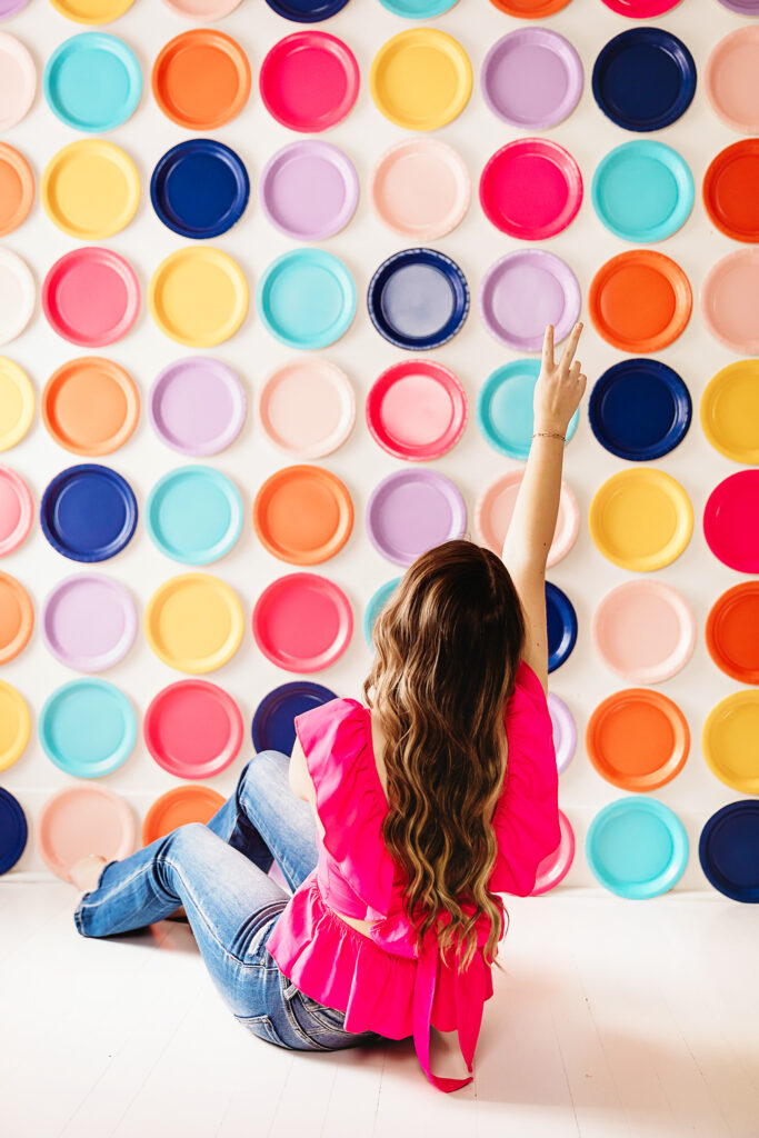 Back-facing peace sign pose in front of rainbow paper plate wall, playful and relaxed