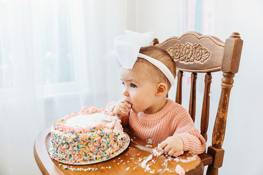 Elsie in pink sweater and white bow, eating sprinkle birthday cake from side view, natural light at Reflection Images by Tracy Rowell in Florence SC