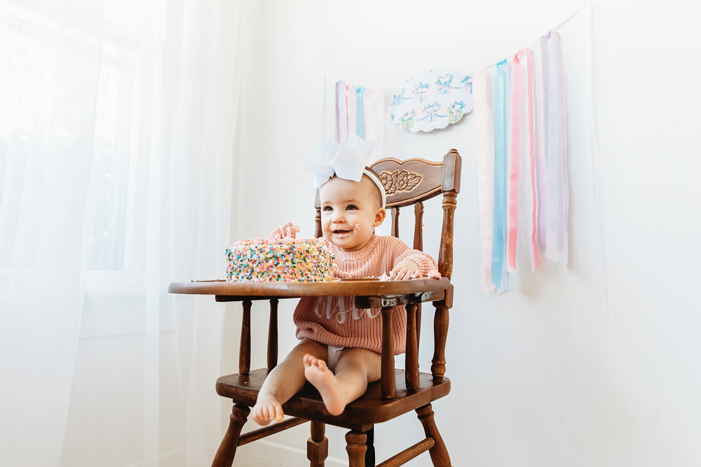Elsie smiling happily in wooden highchair with half-eaten sprinkle cake, birthday backdrop at Reflection Images by Tracy Rowell in Florence SC