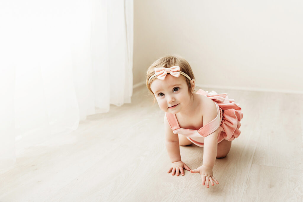 Wide view of Sullivan crawling on the light wooden studio floor in a blush romper and bow headband at Reflection Images by Tracy Rowell in Florence SC.