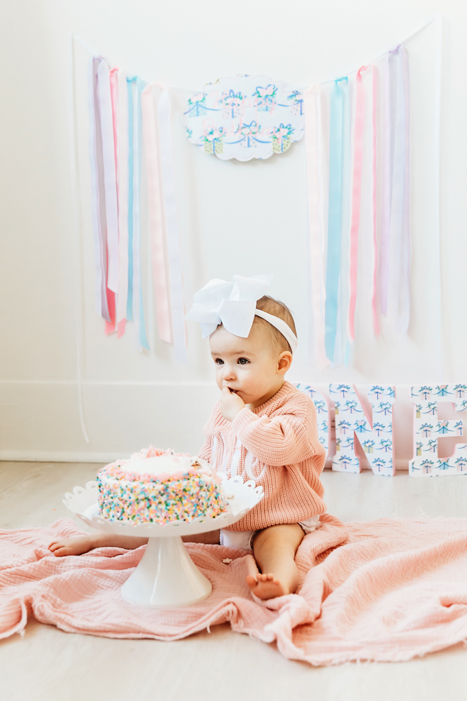Elsie in pink sweater and white bow, sitting on floor beside sprinkle cake, ONE letters and pastel ribbons backdrop at Reflection Images by Tracy Rowell in Florence SC
