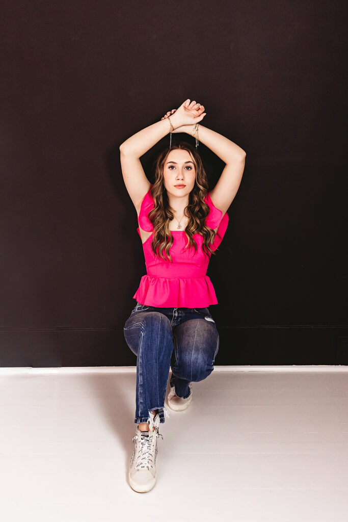 Strong frontal pose against black wall, arms raised, confident and stylish expression