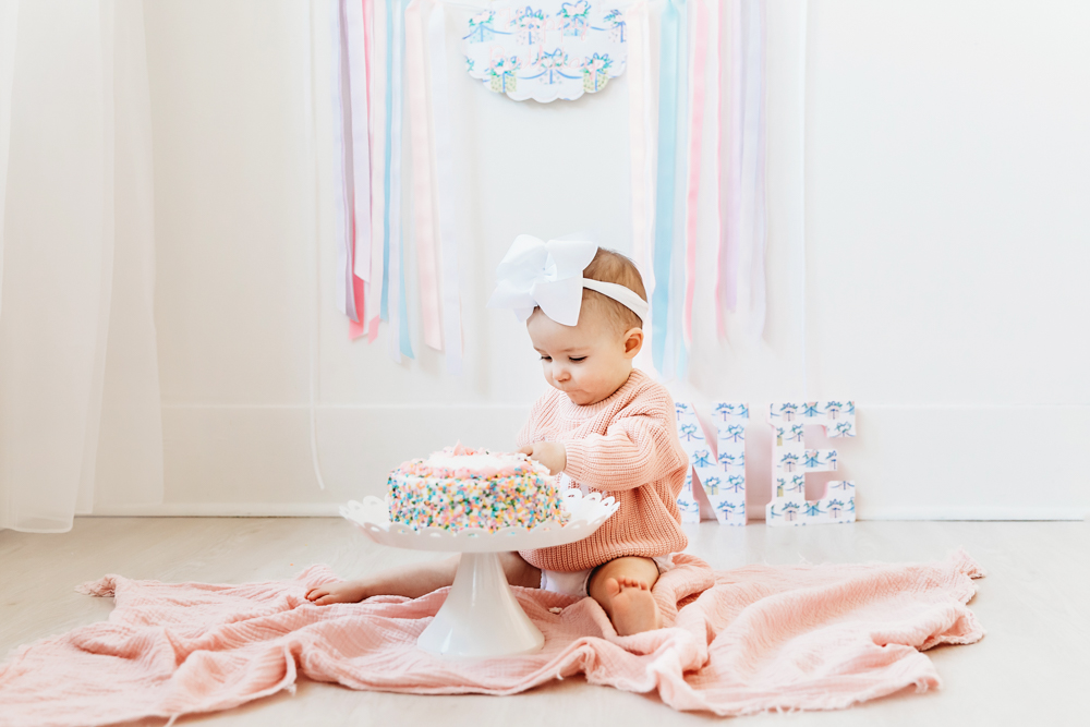 Elsie sitting on pink blanket, poking sprinkle cake with fingers, ONE letters and ribbons backdrop at Reflection Images by Tracy Rowell in Florence SC
