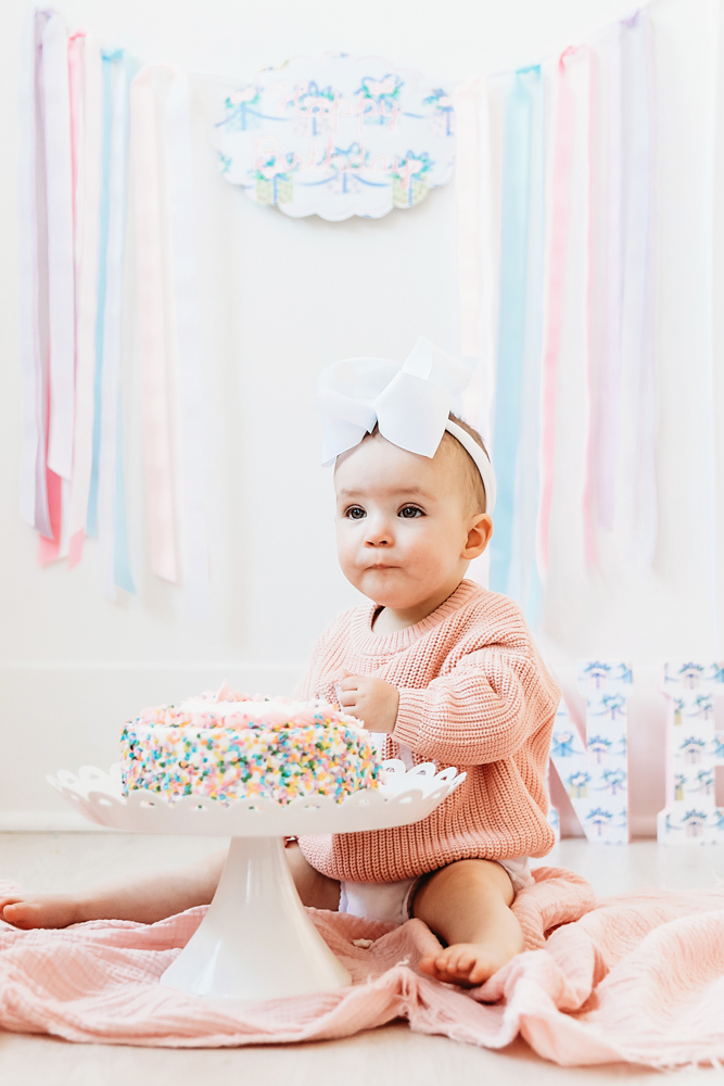 Elsie sitting on pink blanket in white bow and pink sweater, sprinkle smash cake on stand, pastel ribbon backdrop at Reflection Images by Tracy Rowell in Florence SC