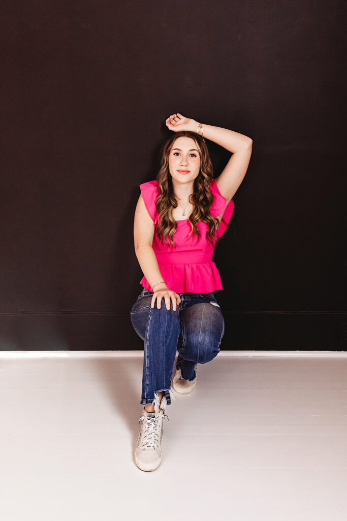 Teen girl kneeling with arm raised, wearing a vibrant pink ruffle top and ripped jeans against a dark backdrop at Reflection Images by Tracy Rowell in Florence SC