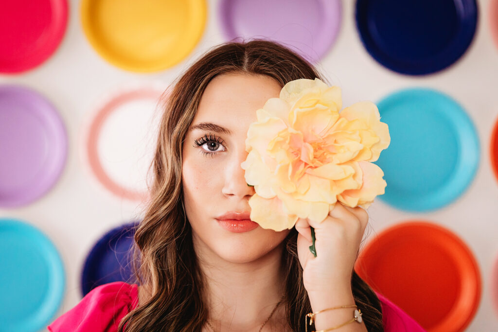 Close-up of teen girl holding yellow flower over one eye with colorful plate wall behind her at Reflection Images by Tracy Rowell in Florence SC