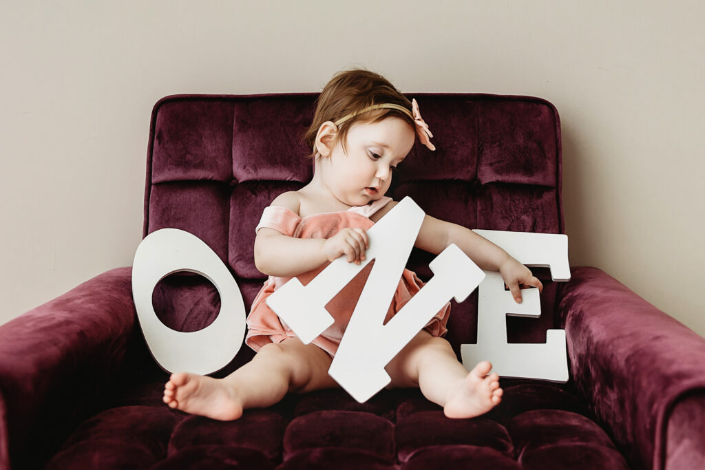 Sullivan lounging across a velvet chair, playfully holding white wooden ONE letters in her lap at Reflection Images by Tracy Rowell in Florence SC.