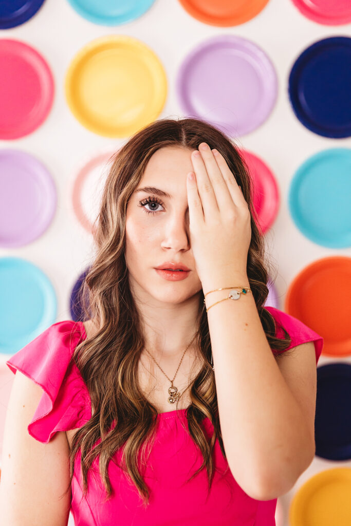 Teen girl sitting on white floor in pink top and jeans, resting her face on hand in front of rainbow plate wall at Reflection Images by Tracy Rowell in Florence SC