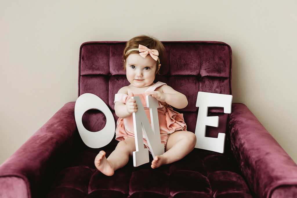 Classic birthday portrait of Sullivan holding up wooden ONE letters while sitting upright on a plum velvet chair at Reflection Images by Tracy Rowell in Florence SC.