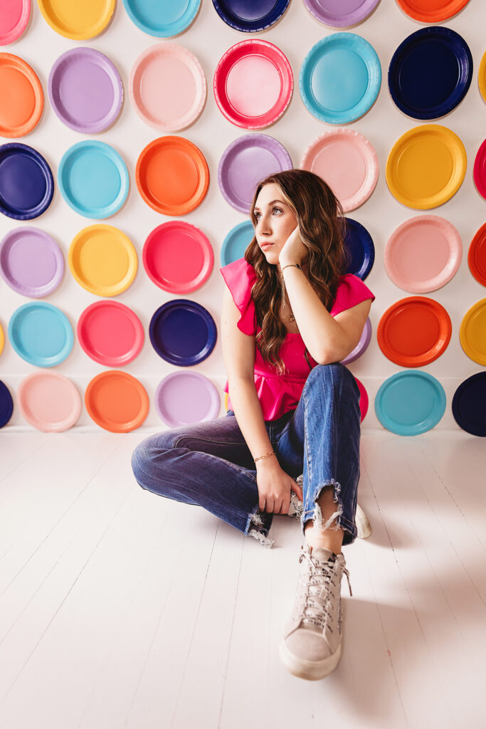 Teen girl laying on her back with long wavy hair spread out, pink top and glitter sneakers, legs up in front of vibrant plate wall at Reflection Images by Tracy Rowell in Florence SC