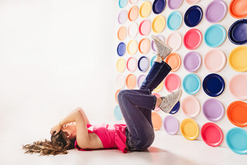 Teen girl smiling while laying sideways on white floor, colorful backdrop of paper plates, pink top and flowing hair at Reflection Images by Tracy Rowell in Florence SC
