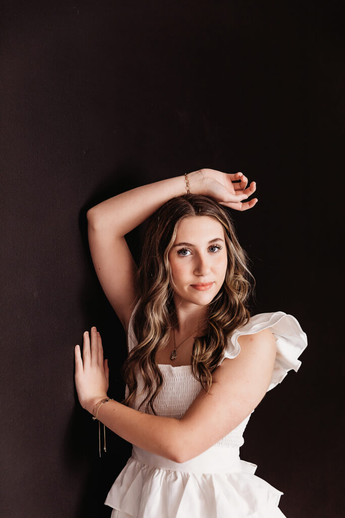 Teen girl standing against black wall in white smocked blouse, arms up and golden bracelet detail at Reflection Images by Tracy Rowell in Florence SC