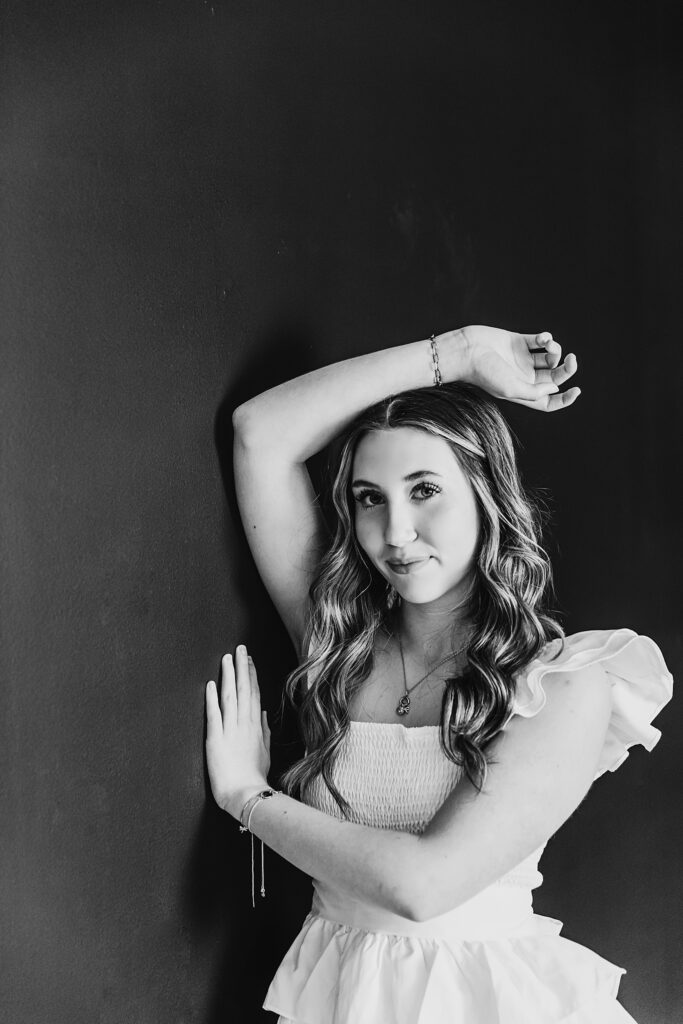 Teen girl standing against black wall in white dress, arms up and golden bracelet detail at Reflection Images by Tracy Rowell in Florence SC